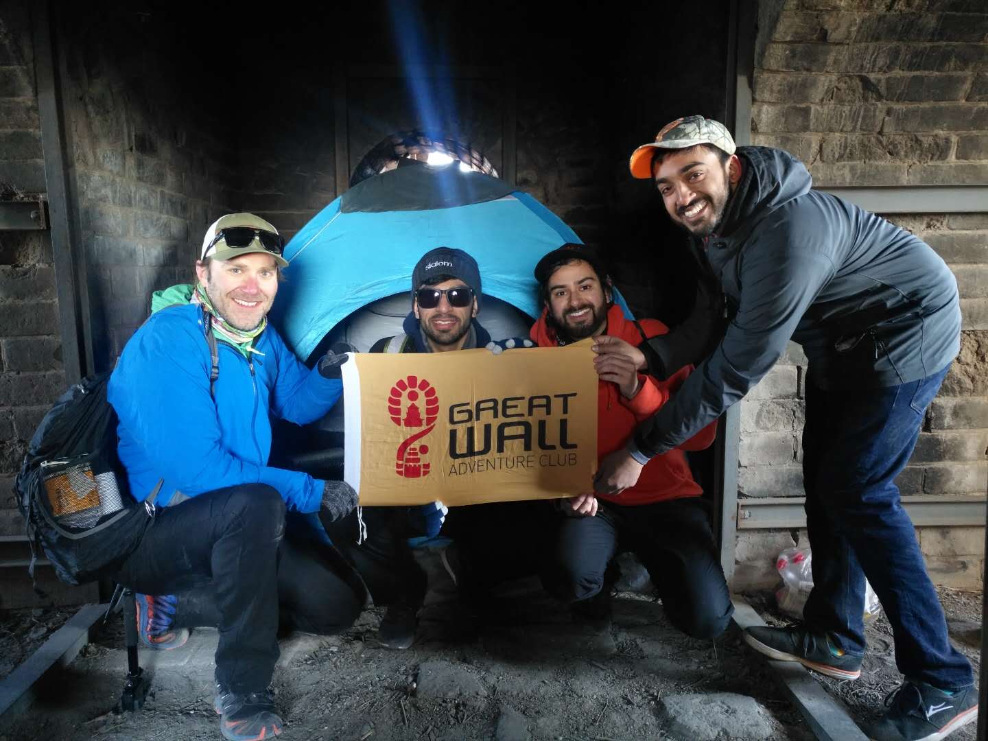 Four travelers holding a Great Wall Adventure Club banner beside a tent inside a brick watchtower