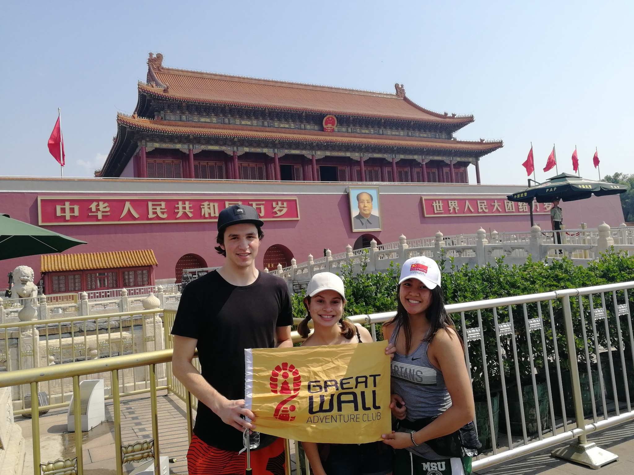 Three travelers holding a Great Wall Adventure Club banner in front of Tiananmen Gate