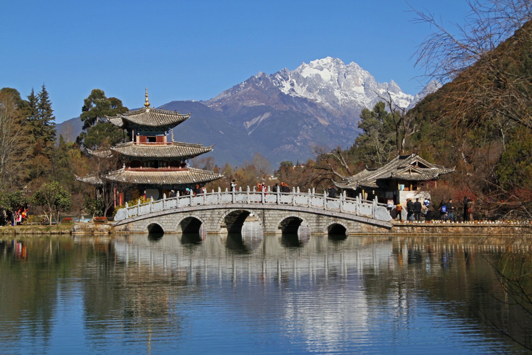 Pavilion and arched stone bridge reflected in a lake beneath snow-covered mountains in Lijiang